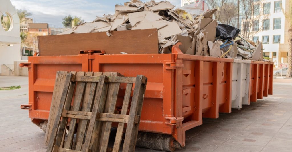A red dumpster is filled above the rim with refuse. There is a commercial building in the background and a dead tree.