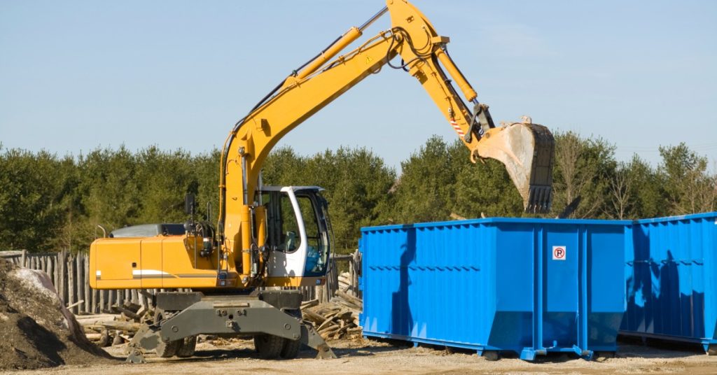 A large excavator is dangling its digging arm above a blue dumpster. There sky is a murky blue color.