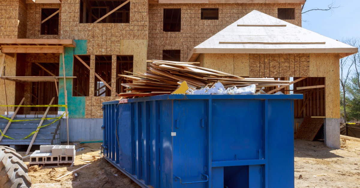 A large blue dumpster is placed outside a residential home that is under construction. There is a tire to the left of the frame.