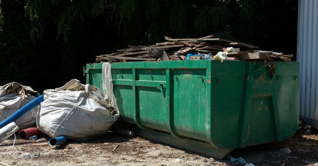 A green dumpster is filled past the brim with garbage. It is in an outdoor environment and there are trees next to it.