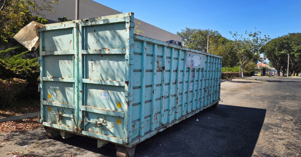 A light blue dumpster is sitting on a road. The blue paint is chipped and reveals rust underneath.