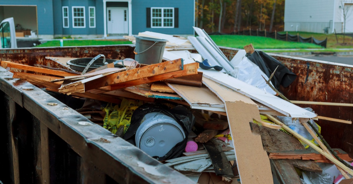 A dumpster is filled with garbage. There is a residential home in the background across the street from the dumpster.
