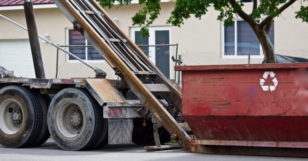 A truck is next to a red dumpster. The bed of the truck is in an upright position, and the dumpster has a recycle icon on it.