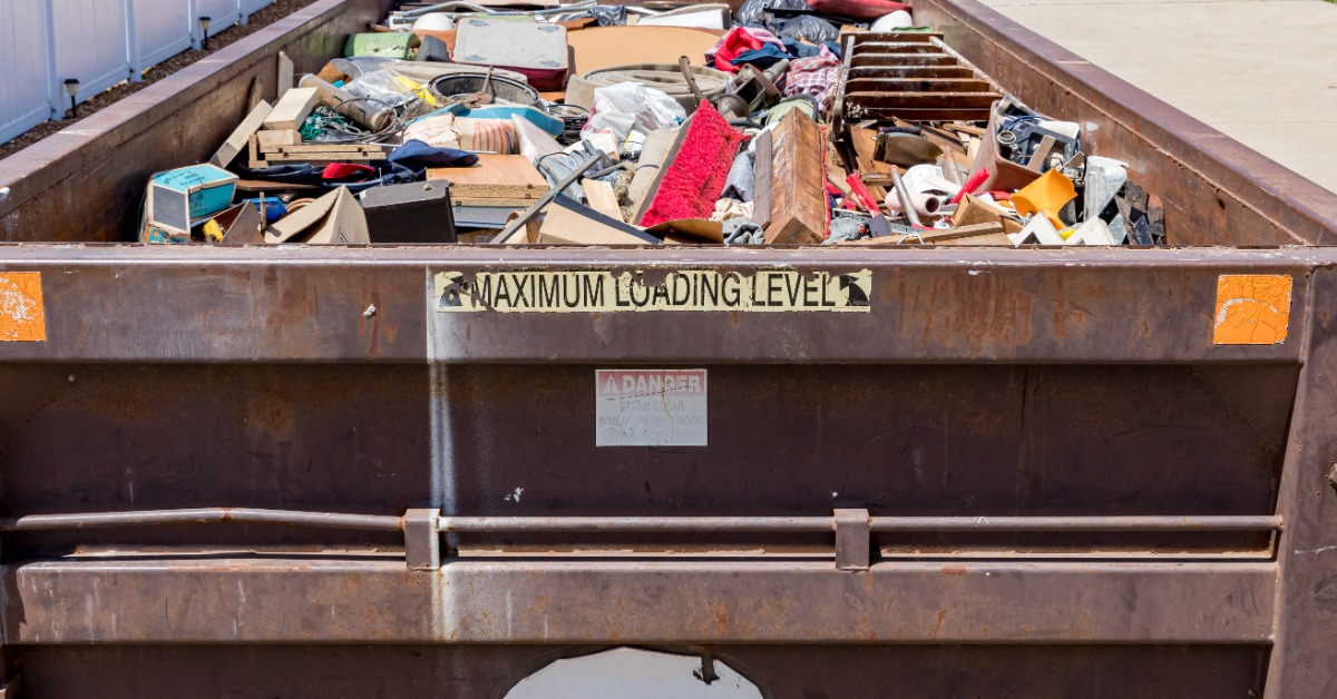 A dumpster with a faded sticker on it is on a driveway. There are mailboxes across the street from the dumpster.